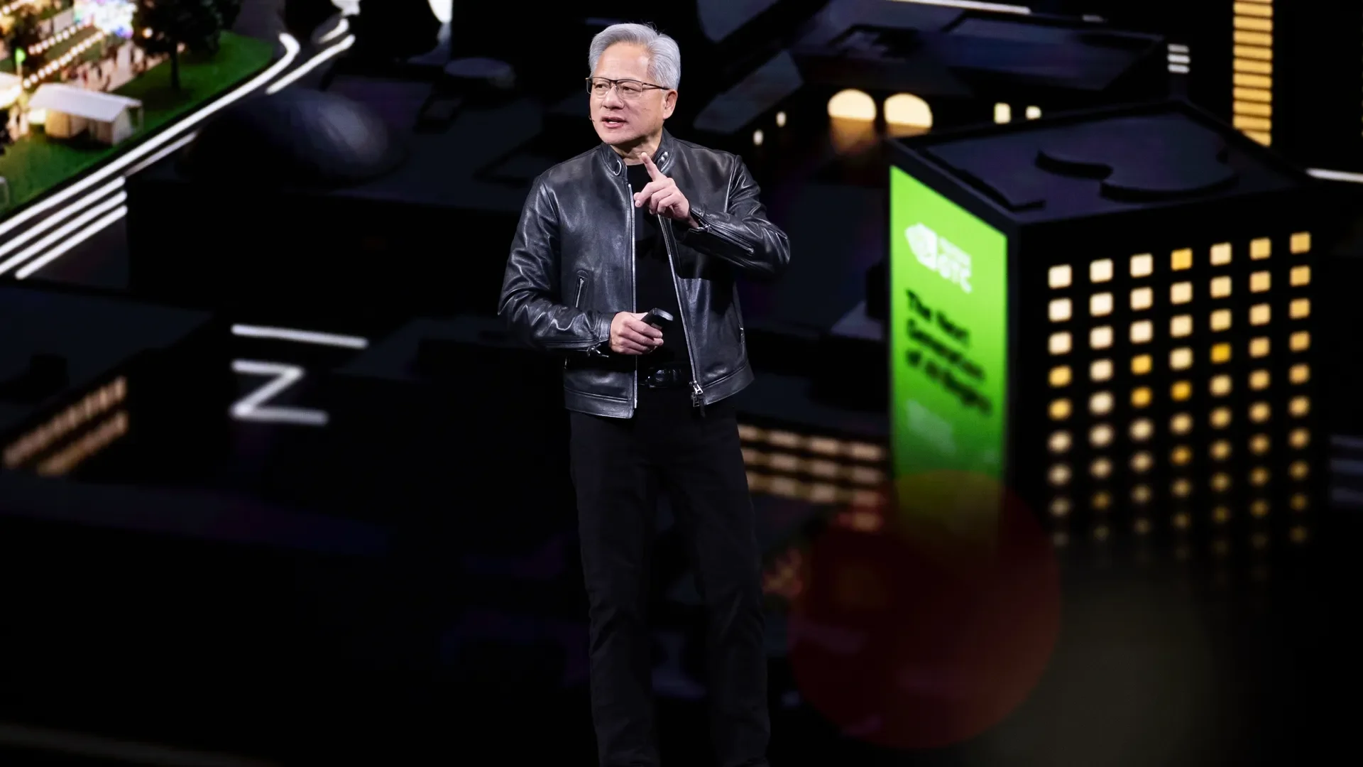 Jensen Huang in a black leather jacket and black pants speaks into a microphone on a dark stage, with a neon cityscape backdrop of lit buildings behind him.